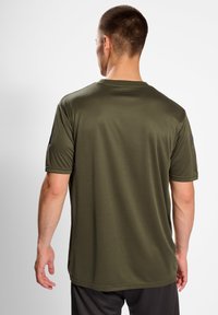 Man med kort hår som står med ryggen mot kameran, klädd i en olivgrön t-shirt med kort ärm och mörka shorts framför en vit bakgrund.