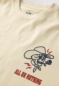 T-shirt couleur crème avec un graphique d'un crâne portant un chapeau et tenant une rose, accompagné d'éclairs rouges et de texte en gras en dessous.