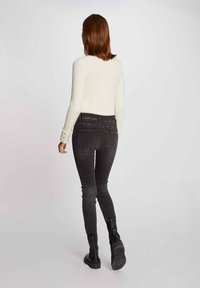 Schwarze skinny Jeans mit hoher Taille, Gesäßtaschen, gelben Nähten, kombiniert mit einem strukturierten cremefarbenen Langarmoberteil. Einfaches Design, figurbetonter Schnitt.