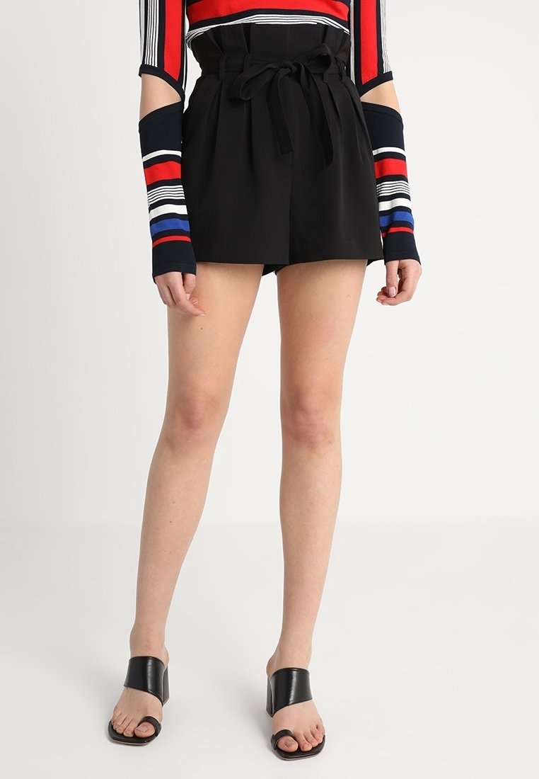 Černé kraťasy s vysokým pasem a opaskem, hladký materiál a volný střih, doplněné o široké černé sandály na klínu.