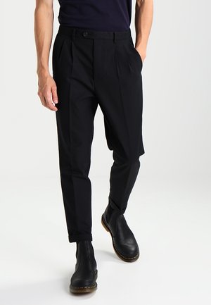 Black trousers with pleats, side pockets, and a straight cut, paired with black ankle boots. Smooth fabric with a tailored design.
