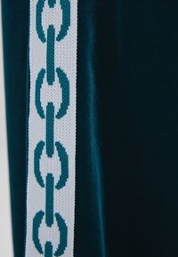 Sametová látka v tmavě tyrkysové barvě s bílou stuhou zdobenou modrým vzorem řetízku po jedné straně. Hladká textura, výrazný barevný kontrast.