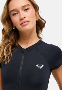 Haut de maillot de bain noir à fermeture éclair à l'avant avec manches courtes, arborant un logo blanc sur la poitrine. Texture lisse, coupe ajustée pour le confort et la liberté de mouvement.