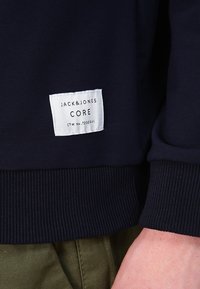 Mörkblå sweatshirt med ribbad mudd och nederkant, försedd med en vit tygetikett som har texten "JACK&JONES CORE" i svart.