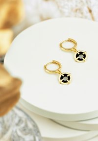 Orecchini a cerchio in oro con un accento nero circolare, caratterizzati da un design geometrico e una texture liscia, esposti su una superficie bianca.