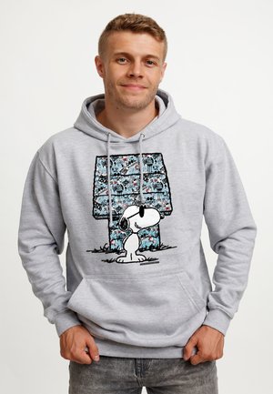 Jeune homme portant un sweat à capuche gris clair avec un graphisme de Snoopy portant des lunettes de soleil devant une lettre bleue à motif "H".