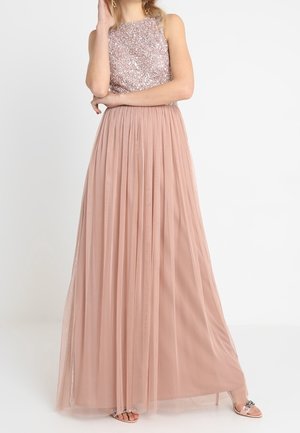 Kobieta ubrana w bezrękawnikową, długą do ziemi suknię w kolorze różu brzoskwiniowego z cekinowym gorsetem i zwiewną spódnicą z tiulu, stojąca z ręką przy twarzy.