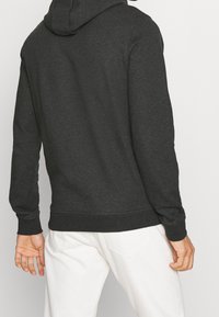 Felpa grigia scura con una texture morbida, dotata di tasca a marsupio, polsini a coste e cappuccio con coulisse, indossata su pantaloni bianchi.