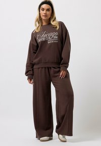 Ruskea huppari, jossa on "Chicago Country Club 1996" -design, yhdistettynä vastaaviin leveisiin housuihin. Pehmeä kangas, rento leikkaus ja neutraalit sävyt.