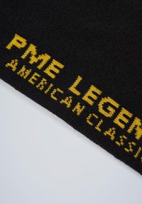 Sort strikket tekstil med gul skrift, der læser "PME LEGEND AMERICAN CLASSIC." Teksturen er blød og lidt fuzzy.