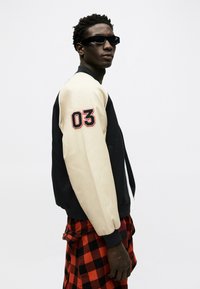 Ung mann iført svarte solbriller, en svart og beige varsityjakke med "03" på ermet, og en rød og svart rutete skjorte knyttet rundt livet.