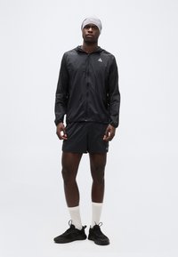 Άνδρας που φοράει μαύρο αθλητικό μπουφάν, μαύρο σορτς, άσπρες κάλτσες μέχρι τον αστράγαλο, μαύρα αθλητικά παπούτσια και γκρι ανοιχτό κορδέλα στο κεφάλι σε απλό λευκό φόντο.