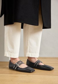 Ballerines en cuir noir à bout pointu, dotées de deux sangles ornées d'œillets en argent. Portées avec un pantalon en velours côtelé crème.