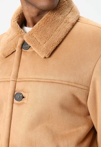 Beige nepbovenjas met een zachte, gestructureerde kraag. Voorzien van twee donkere knopen, een voorpandsluiting en gestikte details.