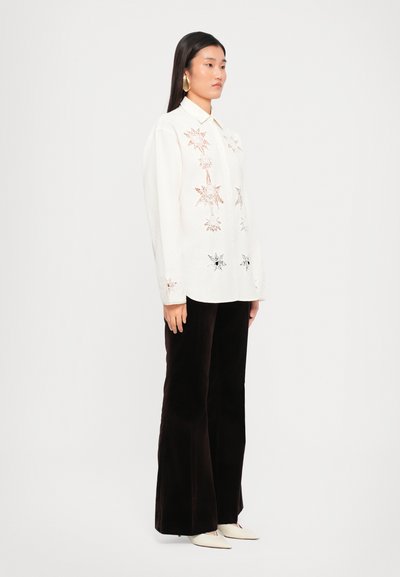 White button-up shirt featuring intricate floral embroidery in black and copper tones, paired with dark velvet flared pants and cream shoes.