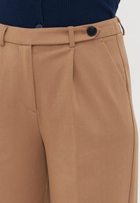 Beige op maat gemaakte broeken met een soepele textuur, voorzien van voorplooien, zijzakken en een zwarte knoopsluiting in de taille.