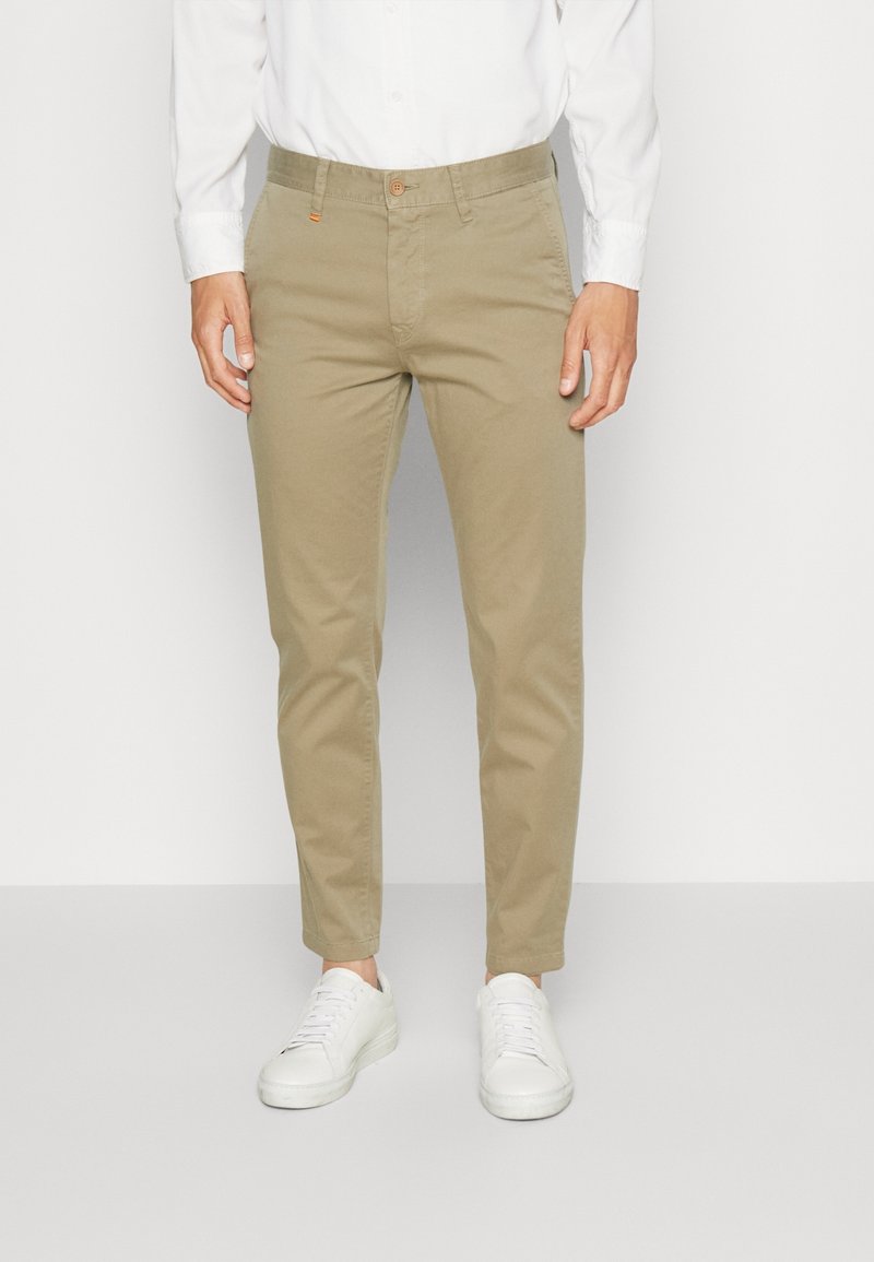 Homme portant un chino beige coupe slim, une chemise blanche à boutons et des baskets blanches, debout devant un fond clair uni.