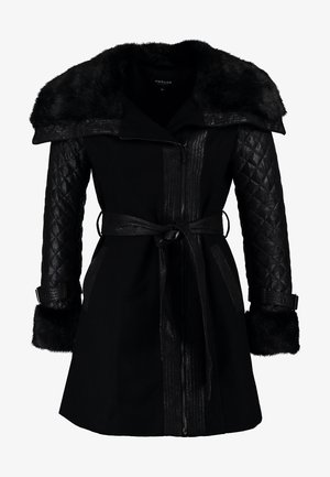 Femme portant un manteau noir bordé de fourrure, un pull gris, un jean bleu, des bottes noires, tenant un grand sac cabas argenté métallique, debout devant un fond uni.