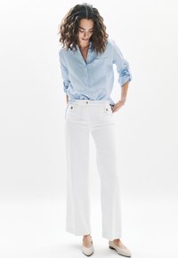 Camicia blu con bottoni e tasca, pantaloni bianchi a gamba larga con tasche frontali e dettagli metallici, e scarpe beige slip-on.