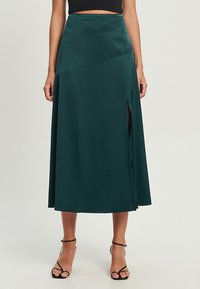 Grüne Satin-Midirock mit hoher Taille, seitlichem Schlitz und drapiertem Design, das eine sanfte Silhouette erzeugt. Getragen mit schwarzen Sandalen mit Riemchen.