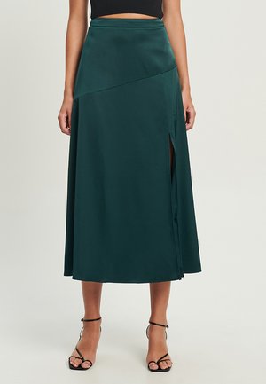 Grüne Satin-Midirock mit hoher Taille, seitlichem Schlitz und drapiertem Design, das eine sanfte Silhouette erzeugt. Getragen mit schwarzen Sandalen mit Riemchen.