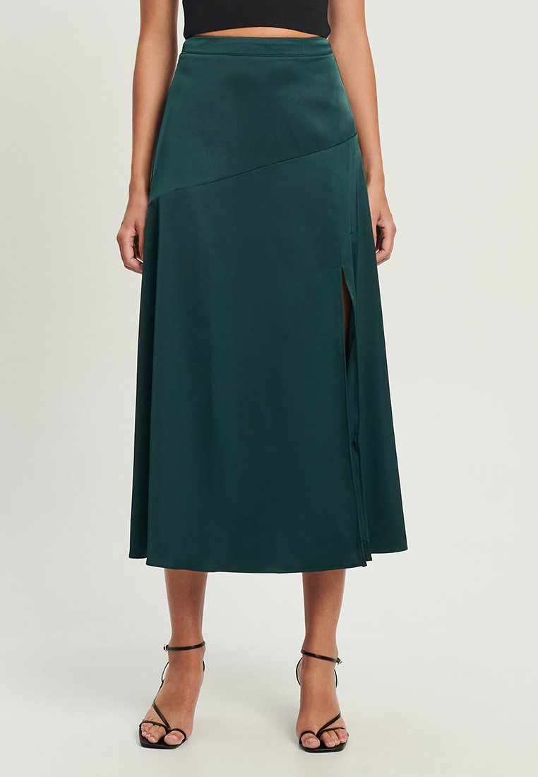 Grüne Satin-Midirock mit hoher Taille, seitlichem Schlitz und drapiertem Design, das eine sanfte Silhouette erzeugt. Getragen mit schwarzen Sandalen mit Riemchen.