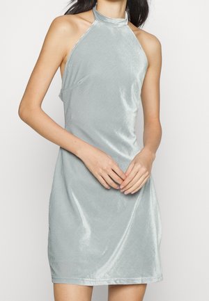 Femme portant une robe en velours argentée sans manches à col halter, mains jointes à la taille, sur fond clair uni.