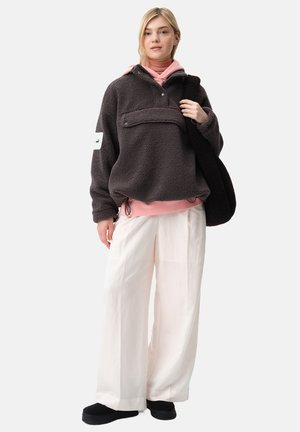 Szary polarowy sweter z kieszenią z przodu, nałożony na różowy golf, zestawiony z luźnymi białymi spodniami i czarnymi butami typu slip-on.