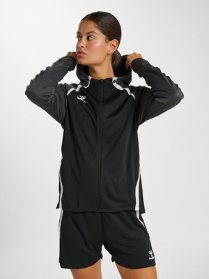 Kobieta poprawia kaptur czarno-szarej sportowej kurtki na zamek, nosząc dopasowane czarne sportowe krótkie spodenki z białymi akcentami.