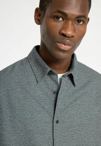 Grijze, gestructureerde shirt met een overhemdkraag, voorzien van een subtiel patroon en twee knopen aan de voorkant, gelaagd over een witte onderhemd.