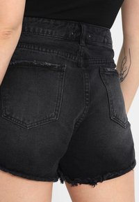 Mustat farkkushortsit, joissa on rispaantunut helma, korkea vyötärö ja kaksi takataskua. Näkyvät saumat ja kulunut tekstuuri korostavat muotoilua.