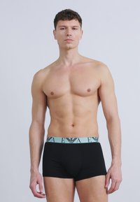 Μυώδης άνδρας που στέκεται αντιμέτωπος, φοράει μαύρα boxer με γαλάζια μέση που φέρει το λογότυπο της μάρκας και γράμματα.