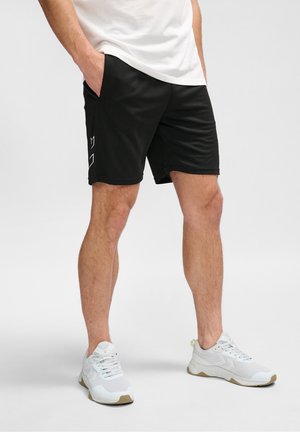 Mand iført sorte shorts med logo på siden, hvide sneakers og hvid t-shirt, stående med hænderne i lommerne på lys baggrund.
