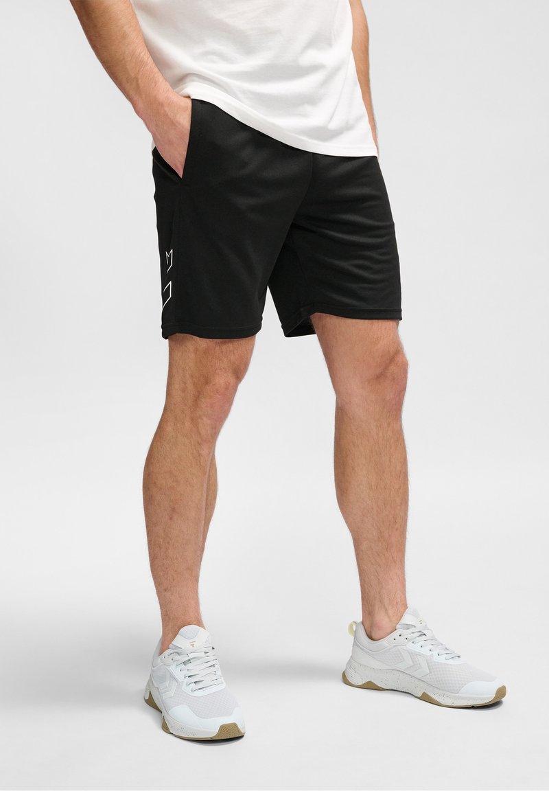 Homme portant un short noir avec un logo sur le côté, des baskets blanches et un t-shirt blanc, debout les mains dans les poches sur un fond clair.