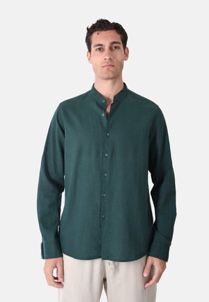 Uomo che indossa una camicia verde scuro con colletto mandarino abbottonata e pantaloni beige chiaro, in piedi davanti a uno sfondo bianco uniforme.