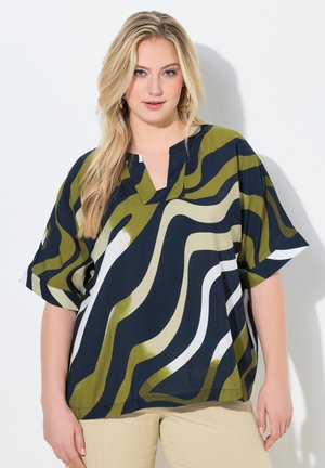 Blondynka w luźnej koszuli z krótkim rękawem z zielonymi, granatowymi i białymi abstrakcyjnymi falistymi wzorami oraz beżowymi spodniami, stojąca na białym tle.