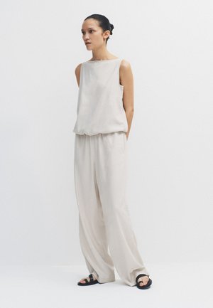 Kvinde med mørkt hår bundet tilbage iført løs, ærmeløs, lys beige top og bukser med brede ben, stående med hænderne bag ryggen i en neutral setting.