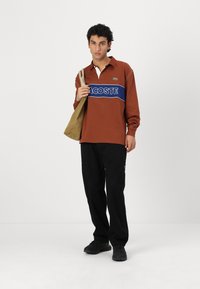 Hnedý dlhý rukáv polo s modrým pruhom a logom "LACOSTE", kombinovaný s čiernymi nohavicami, čiernymi topánkami a béžovou ramennou taškou. Detail bieleho goliera.