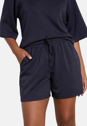 Persoon draagt donkerblauwe casual korte broek met elastische tailleband en trekkoord, gecombineerd met een bijpassend los vallend T-shirt met korte mouwen, hand in de zak.