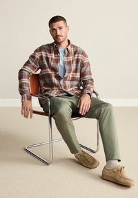 Camicia in flanella a motivi plaid nei toni ruggine e grigi, t-shirt azzurro chiaro, pantaloni verde oliva, scarpe in suede, seduto su una sedia moderna in metallo.