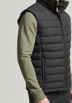 Mand iført sort polstret vest over olivengrøn langærmet skjorte, vendt mod højre med delvist synlig overkrop og arm.