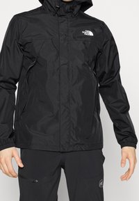 Black waterproof jacket with hood, zippered pockets, and The North Face logo on chest, worn with black pants with zippered pocket.