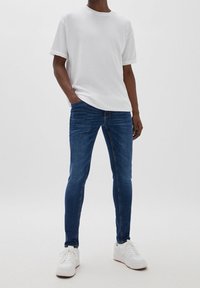 Man som bär en enkel vit t-shirt, slim fit mörkblå jeans och vita sneakers, står med en hand i fickan mot en enfärgad bakgrund.