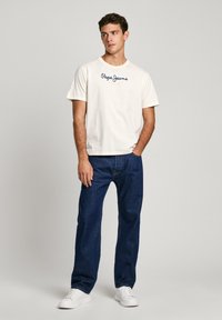 Balta īsām piedurknēm T-krekls ar zilu "Pepe Jeans London" tekstu, kopā ar tumši zilu džinsu biksēm un baltām sporta kurpēm. Kopumā vienkārša dizaina.