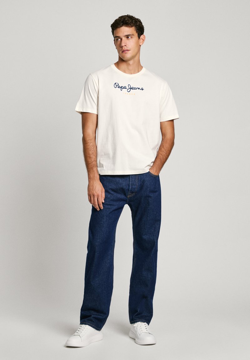 Balta īsām piedurknēm T-krekls ar zilu "Pepe Jeans London" tekstu, kopā ar tumši zilu džinsu biksēm un baltām sporta kurpēm. Kopumā vienkārša dizaina.