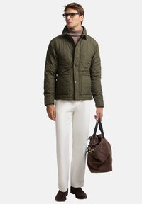 Giacca trapuntata verde con collo a camicia, due tasche frontali, abbinata a pantaloni crema e scarpe scamosciate marroni, con una borsa marrone in mano.