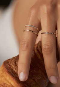 Tre anelli su una mano: una fede d'oro, una fede con accenti di diamanti e una sottile fede in oro rosa, poggiati su un croissant con superficie texturizzata.