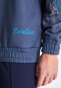 Marineblå sweatshirt med broderet "Carlo Colucci"-logot. Har elastiske manchetter og et struktureret stof med farverige abstrakte mønstre på ærmerne.