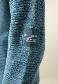 Strukturierter blauer Strickstoff mit einem eingewebten Etikett, das "LDN", "MCMLXXXII" und Koordinaten zeigt. Der Stoff hat ein gleichmäßiges, geripptes Muster.