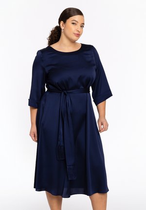 Navy satijnen jurk met driekwart mouwen, getailleerde taille en riem met kwastaccenten, vloeiende rok die onder de knie valt.
